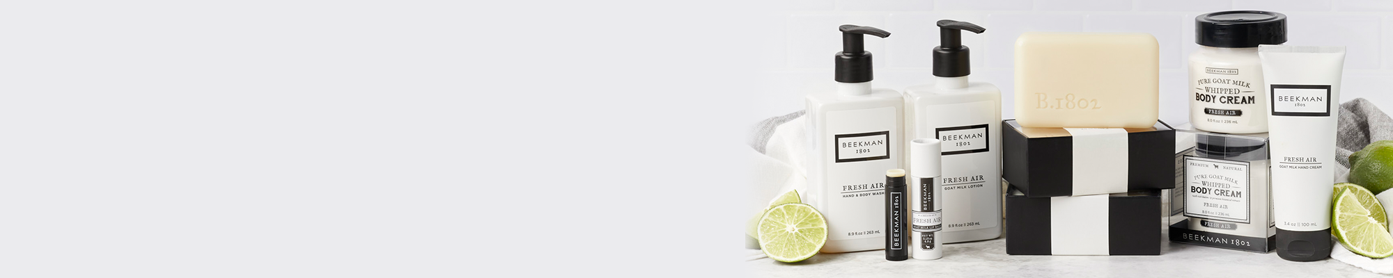 Products from Beekman 1802's Fresh Air collection all displayed together showing the bar soaps, body cream, hand & Body wash, hand cream, lotion and lip balm to the far right of the screen, surrounded by limes on a light gray tile background.  