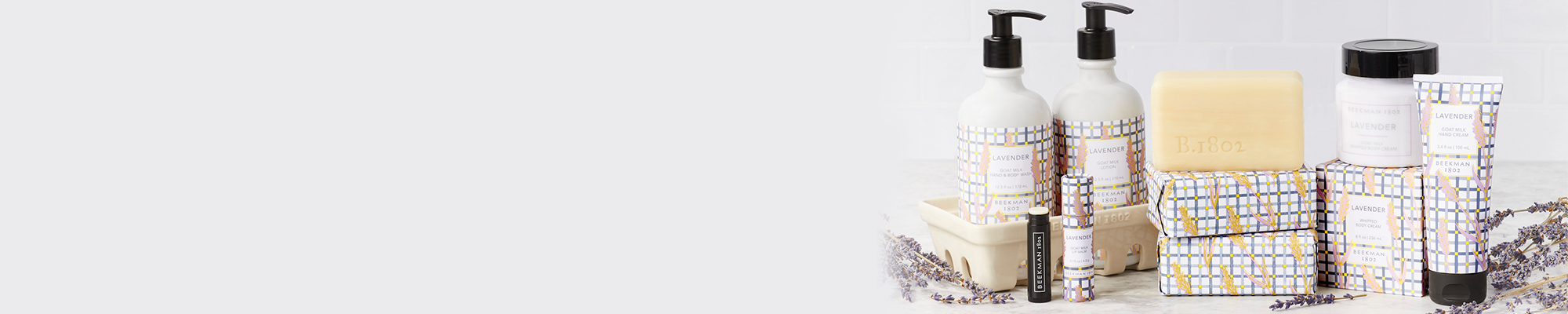 Products from Beekman 1802's Lavender Collection which includes the caddy set, lip balm, bar soap, body cream and hand cream all displayed next to each other on the far right side of the screen, surrounded by lavender flowers on a light grey tile background. 