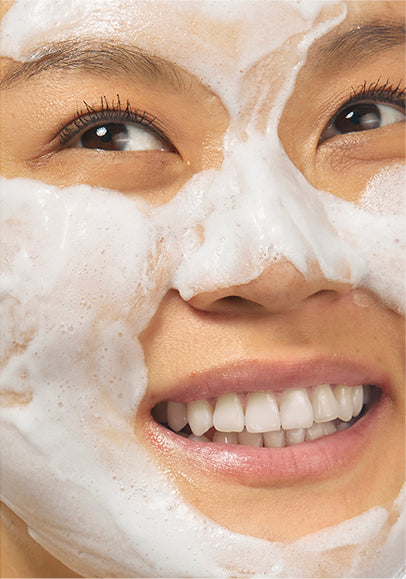 Up close shot of model smiling with her teeth and looking off to the right, while wearing the Beekman 1802 Milk Foam calming Bubble mask on her face, showing all the bubbles that have formed on her skin. 