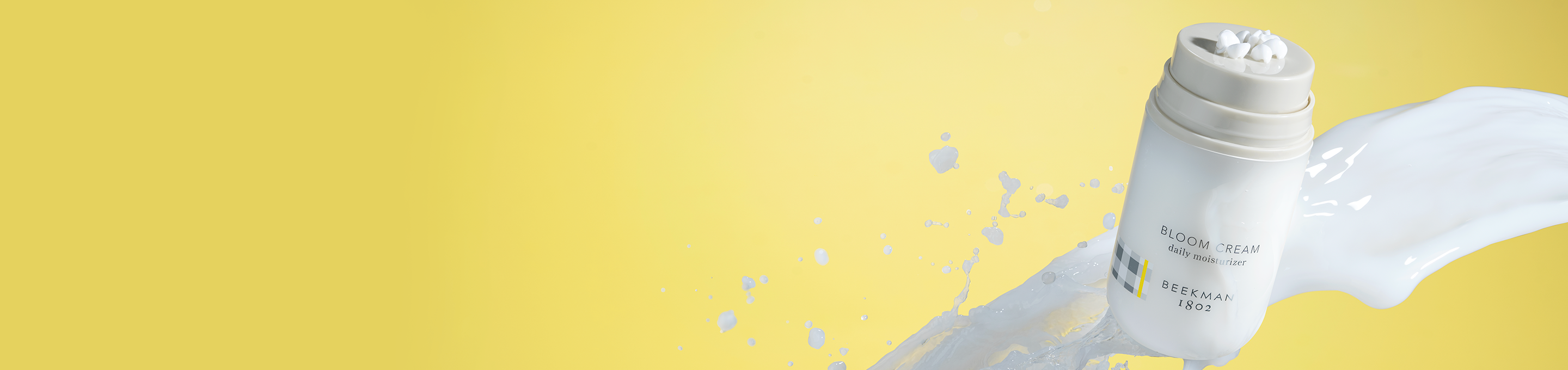 Bottle of Beekman 1802's Bloom Cream Daily Moisturizer mid-air, surrounded by a milky splash, on a yellow background to the far right of the screen.